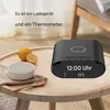 A black digital alarm clock with a wireless charging pad on top, placed on a wooden table next to a bowl and lemons. Produktbild-Vorschau
