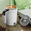 A white self-stirring mug with a digital temperature display is shown on a wooden table, with a lid next to it. Coffee beans are scattered in the foreground, and a green pillow is in the background. Produktbild-Vorschau