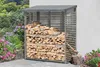 A wooden log store filled with firewood, placed outdoors next to a white wall, with a garden and flowers in the background. Produktbild-Vorschau