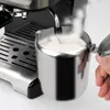 Close-up of a person's hand holding a milk frothing pitcher next to an espresso machine. Produktbild-Vorschau