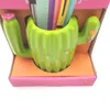 A green ceramic cactus-shaped pen holder with pink and orange floral accents, holding colorful pens and a ruler, is displayed in its packaging. Voir les images du produit