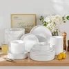 A collection of white ceramic dishes and bowls are arranged on a wooden surface, with a framed picture and a bouquet of white flowers in the background. Produktbild-Vorschau