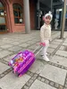 A young child is pulling a suitcase on a paved walkway outside a building. Anteprima dell’immagine prodotto