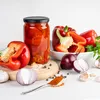 A still life arrangement of red bell peppers, some whole, some cut, a jar of preserved peppers, red onions, garlic, peppercorns, and paprika on a white surface with a wooden cutting board. Podgląd zdjęcia produktu