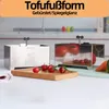 Two stainless steel tofu presses are shown on a kitchen counter, with tomatoes and other vegetables in the foreground and background. Produktbild-Vorschau
