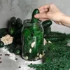 A hand is placing a cucumber into a glass jar filled with other cucumbers, dill, and garlic. The jar is surrounded by more cucumbers, dill, garlic, peppercorns, and salt. Produktbild-Vorschau
