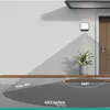 An outdoor wall lamp with a motion sensor is shown mounted on a white wall next to a door, illuminating a pathway and a potted plant. Diagrams indicate its coverage range and height. Produktbild-Vorschau