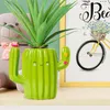A green ceramic cactus-shaped pen holder with a smiling face, holding a plant, sits on a shelf next to a teddy bear and a framed picture of a bicycle. Voir les images du produit