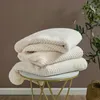A stack of three cream-colored blankets on a small, round table with gold legs, next to a plant. Produktbild-Vorschau