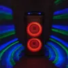 A tall, black speaker with two red illuminated speakers is shown in a dark room with colorful light effects projected onto the sides. Voir les images du produit