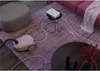 A pink vacuum cleaner is shown on a purple rug in a living room setting, with a small table and a sofa visible. Produktbild-Vorschau