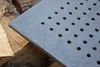 A close-up of a square cast iron grate with many small holes, resting on a pile of firewood. Produktbild-Vorschau