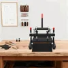 A manual leather cutting and embossing machine is displayed on a wooden table, with various leatherworking tools and materials arranged around it. A diagram of leather patterns hangs on the wall in the background. Náhled obrázku produktu