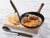 A frying pan with food, bread, and cooking utensils on a wooden board. Voir les images du produit