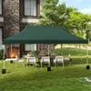 A green canopy tent is set up in a backyard with a table and chairs underneath, next to a house with a fireplace. Produktbild-Vorschau
