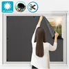 A woman is applying a dark gray blackout film to a window from the inside. Produktbild-Vorschau
