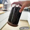 Hand, die ein klares Glas-Vorratsglas mit Holzdeckel hält, gefüllt mit dunkel gerösteten Kaffeebohnen, in einer modernen Küche Produktbild-Vorschau