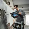 A man is using a Bosch rotary hammer to break tiles off a wall while standing on a ladder. Produktbild-Vorschau