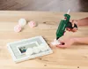 A person is using a hot glue gun to attach a pink rose to a picture frame decorated with white roses. Produktbild-Vorschau