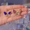 Zwei goldene Ketten mit Schmetterlingsanhängern, einer mit blauen Flügeln und der andere mit rosa und klaren schillernden Flügeln, in einer Hand gehalten Náhľad obrázku produktu