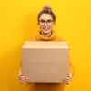 A woman holding a cardboard box in front of a yellow background. Produktbild-Vorschau