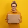 A woman wearing a yellow sweater and glasses holds a brown cardboard box in front of a yellow background. Produktbild-Vorschau