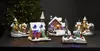 A collection of illuminated Christmas village houses and churches, set against a dark background with a blurred Christmas tree and lights. Podgląd zdjęcia produktu