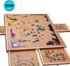 A puzzle board with many puzzle pieces spread across it and in drawers. The board is brown and has a wooden frame. There are also drawers that slide out from the sides and bottom, containing more puzzle pieces. Produktbild-Vorschau