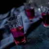 Several shot glasses filled with a purple liquid are arranged on a dark blue cloth. Náhled obrázku produktu
