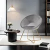 A modern living room scene with a gray velvet accent chair, a small round coffee table with a vase of flowers and a teacup, and a bookshelf in the background. Produktbild-Vorschau