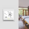 A smart thermostat mounted on a white wall, with a blurred background of a hotel room with two beds and large windows. Náhled obrázku produktu