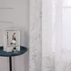 A close-up of a white sheer curtain with a gray branch pattern, next to a small round table with books and a diffuser. Produktbild-Vorschau