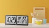 A digital clock with temperature and humidity display sits on a shelf next to kitchenware. Produktbild-Vorschau