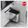 A stainless steel trash can with a black lid is pulled out from a kitchen cabinet. Náhled obrázku produktu