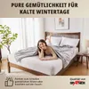 A woman is lying on a bed covered by a blanket, smiling at the camera. Produktbild-Vorschau