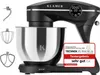A black stand mixer with a silver bowl and several attachments is displayed against a white background. A red and white badge with text is affixed to the front of the mixer. Produktbild-Vorschau