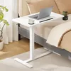 A white adjustable height table with a laptop, cup, and drink holder is placed next to a bed with tan bedding. Produktbild-Vorschau