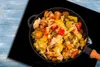 A close-up overhead shot of a black frying pan filled with a colorful mix of cooked vegetables, placed on a black stovetop with a blue wooden background. Náhľad obrázku produktu
