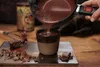 Hot chocolate being poured from a pot into a mug, with chocolate pieces and a whisk on a wooden surface. Produktbild-Vorschau