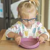 A young child wearing glasses and a colorful shirt is sitting at a wooden table, holding a pink suction plate. Náhľad obrázku produktu