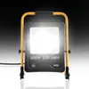 A bright LED work light with a yellow frame and handle, shown against a dark, gradient background. Voir les images du produit