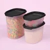 Three clear plastic food storage containers with black lids are stacked and arranged on a light pink background. The largest container on the left is filled with colorful cereal loops. The middle container is stacked on top of the bottom container on the r Produktbild-Vorschau