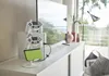A white controller stand with two white game controllers and a green handheld gaming device is placed on a white dresser in front of a large window with a view of greenery. Produktbild-Vorschau