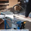 A person is using a table saw in a workshop setting, cutting a piece of wood. There are stacks of wood in the background. Anteprima dell’immagine prodotto