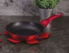 A red frying pan with a speckled interior sits on a dark countertop, next to a potted plant. Náhled obrázku produktu