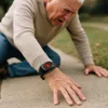 An elderly man is on the ground, reaching out with his hand, wearing a black SOS device on his wrist. Náhľad obrázku produktu