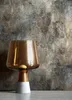 A modern table lamp with a glass shade and a concrete base is placed on a surface against a textured wall. Produktbild-Vorschau