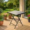 A square folding table with a wood-grain effect top stands on a wooden deck in a garden setting. Potted plants and greenery surround the table, with a house in the background. Produktbild-Vorschau