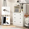 A white dresser with three drawers and an open shelf above, decorated with a small speaker, a framed picture, and a vase with flowers. Two inset images show an anti-tip bracket and a diagram illustrating its use. Podgląd zdjęcia produktu