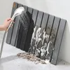 A hand holding a shower head sprays water onto a gray slatted bath mat, creating foam and washing away dirt. Anteprima dell’immagine prodotto